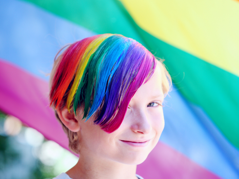 criança com cabelo nas cores do arco-íris e ao fundo uma bandeira lgbtqia+