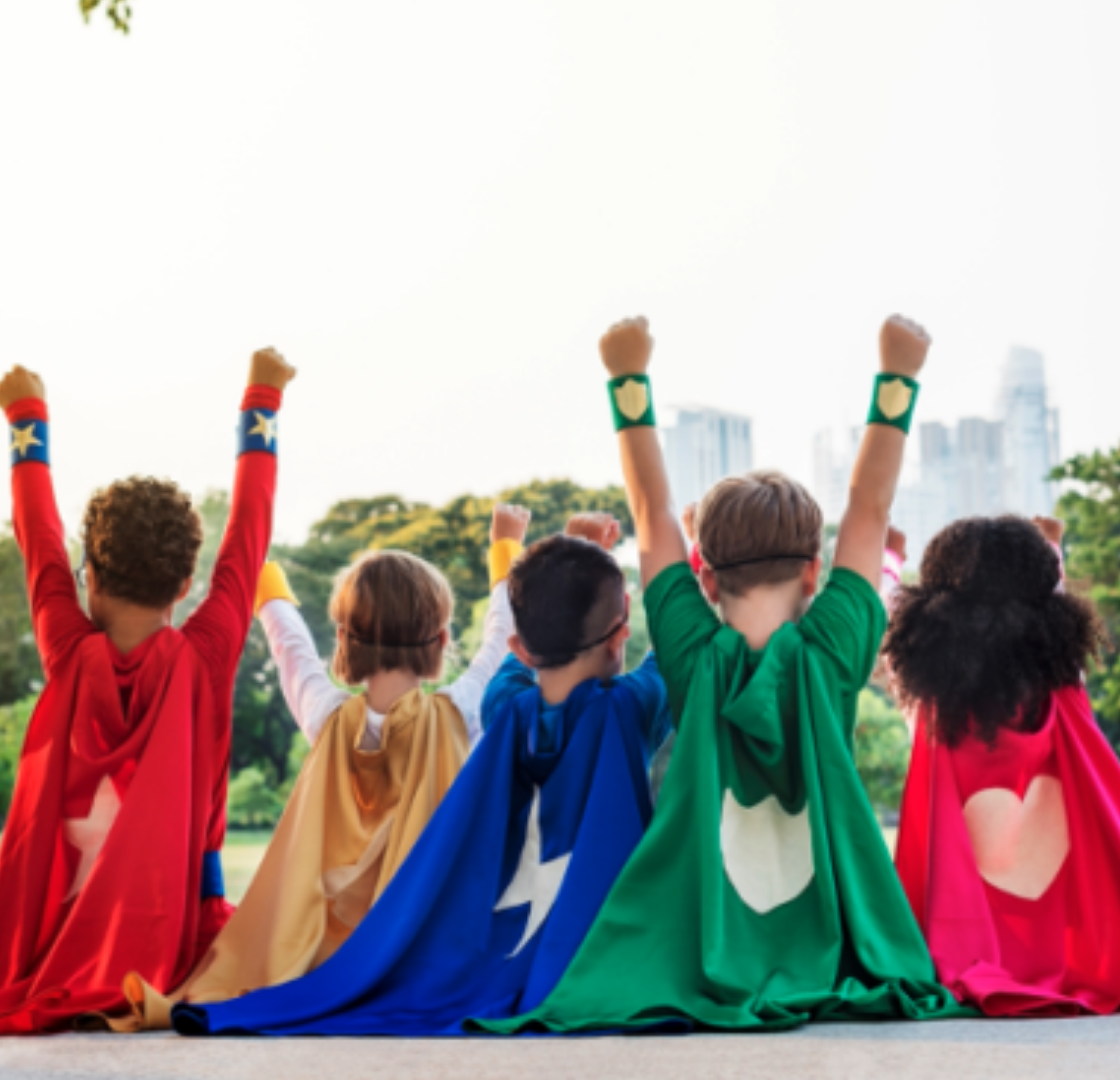 Foto de 5 crianças, com características físicas distintas, sentadas de costas, lado a lado. Todas usam uma capa de super herói, cada uma de uma cor e estão com os braços para cima e com os punhos fechados.