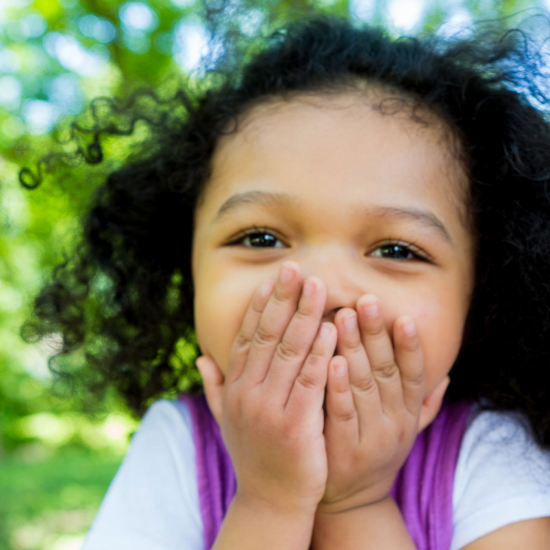 Foto de rosto de uma menina de pele parda e cabelos pretos crespos com as mãos na boca olhando pra frente com ar de surpresa.
